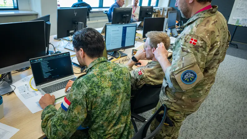Twee Deense militairen, van wie een staand met een Nederlandse collega naar twee beeldschermen kijkt. 