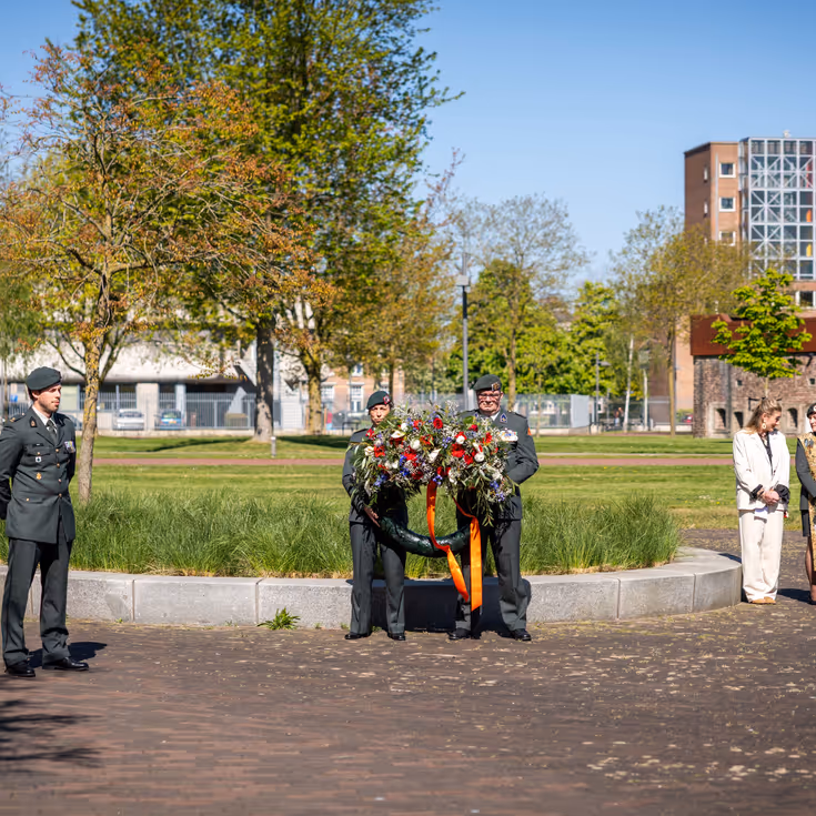Twee militairen houden een bloemenkrans vast onder een strakblauwe lucht. 