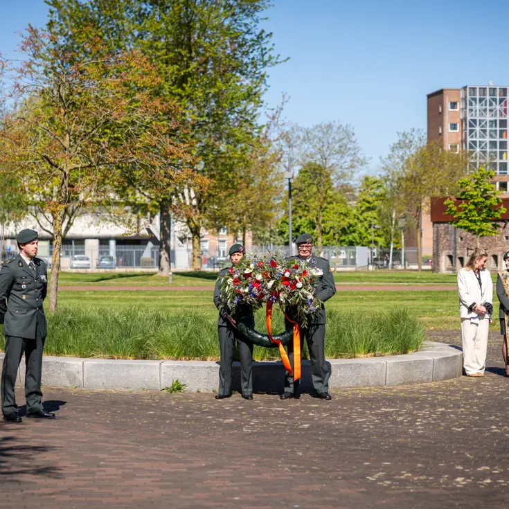 Twee militairen houden een bloemenkrans vast onder een strakblauwe lucht. 