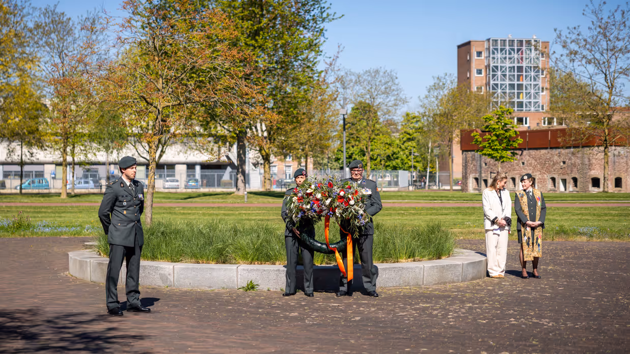 Twee militairen houden een bloemenkrans vast onder een strakblauwe lucht. 