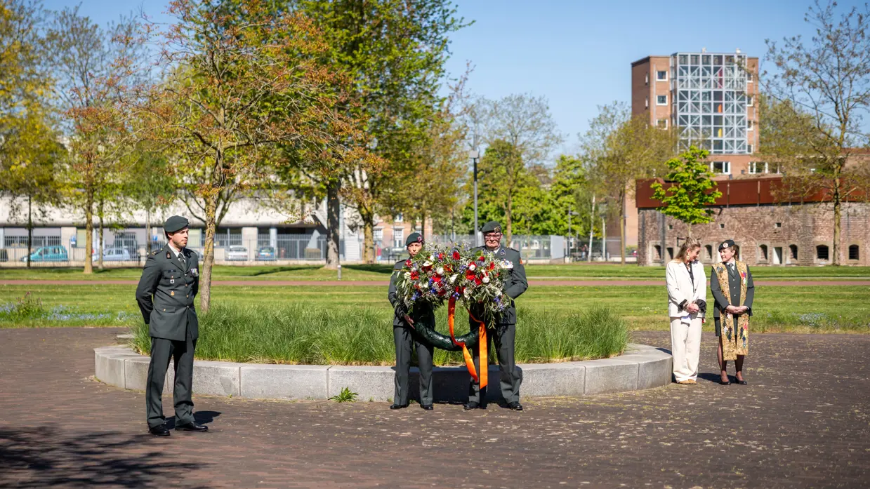 Twee militairen houden een bloemenkrans vast onder een strakblauwe lucht. 
