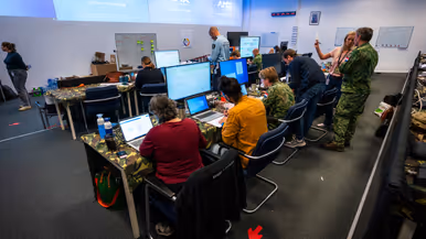 Drie burgermedewerkers en militairen zitten in een grote ruimte achter beeldschermen met aan de wand enkele heel grote beeldschermen.