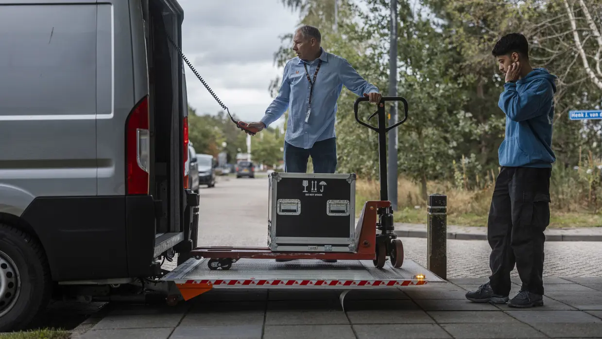 Twee medewerkers laden een flightcase met ICT-apparatuur uit een bestelbus voor de opbouw van Falcon Leap.