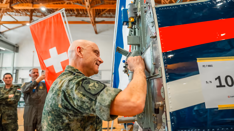 Een militair zet zijn handtekening op de romp van een lesvliegtuig, bijgestaan door andere genodigden.