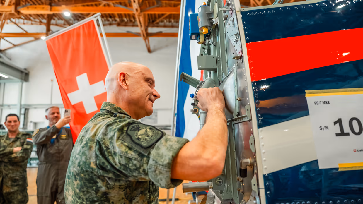 Een militair zet zijn handtekening op de romp van een lesvliegtuig, bijgestaan door andere genodigden.