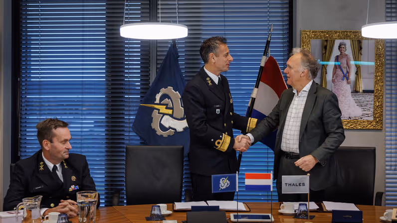 Twee mannen schudden elkaar de hand, van wie één in militair tenue. Ze staan achter een tafel. Op de achtergrond hangt een schilderij van de koningin en staan vlaggen, die ook in het klein op het bureau staan.