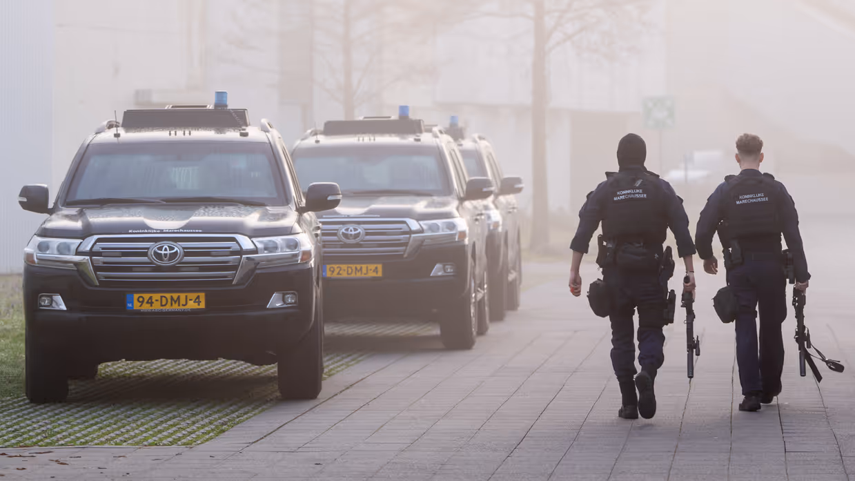 Twee marechaussees lopen op een betegeld pad. Ze dragen donkerblauwe uniformen met "Koninklijke Marechaussee" op de rug en zijn bewapend. Achter hen rijden drie zwarte, zware terreinwagens van de Koninklijke Marechaussee
