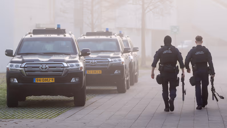 Twee marechaussees lopen op een betegeld pad. Ze dragen donkerblauwe uniformen met "Koninklijke Marechaussee" op de rug en zijn bewapend. Achter hen rijden drie zwarte, zware terreinwagens van de Koninklijke Marechaussee