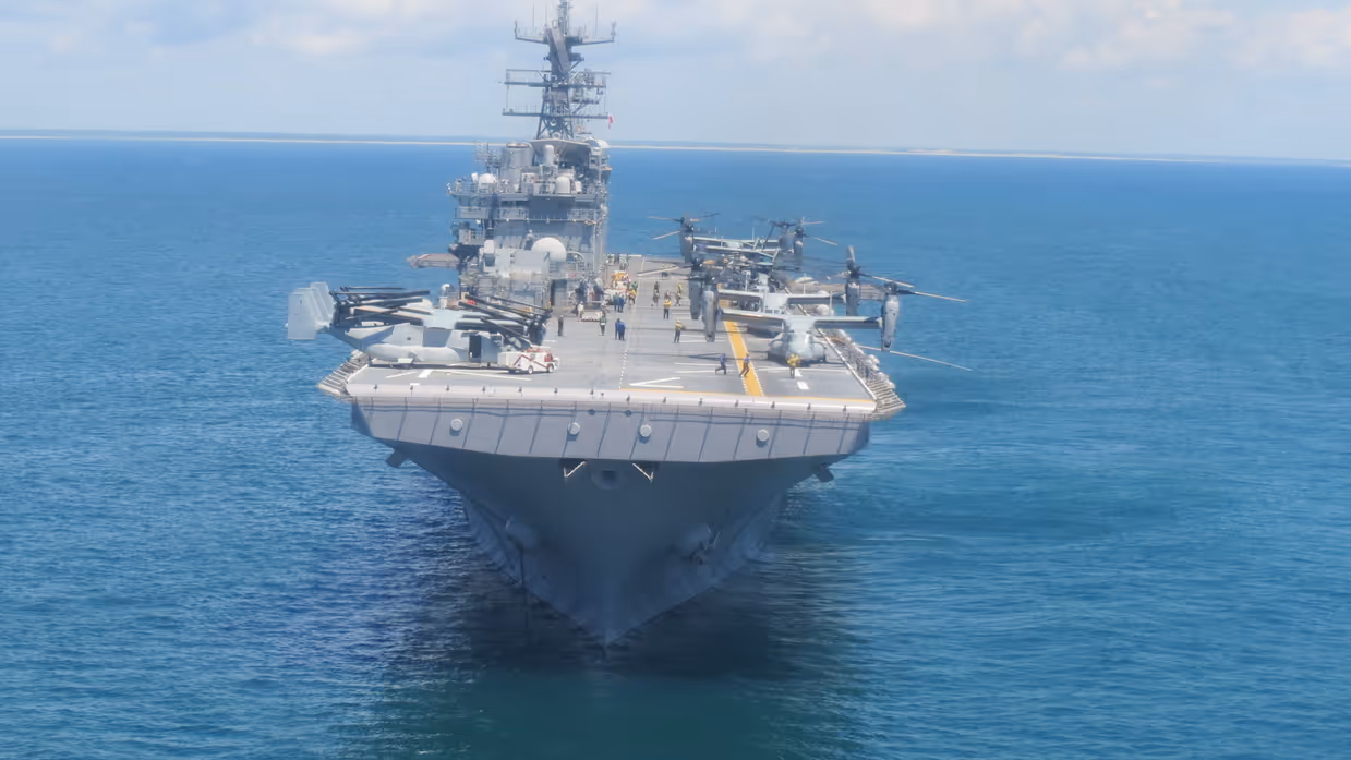 Een groot grijs oorlogsschip vaart op rustig blauw water, met helikopters en vliegtuigen op het dek, onder een lichtbewolkte lucht.