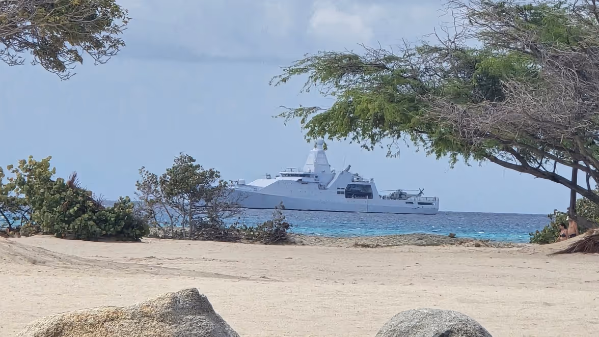 Een rustig strand met grote grijze stenen, bomen, en een wit oorlogsschip op de kalme zee in de verte onder een licht bewolkte lucht.