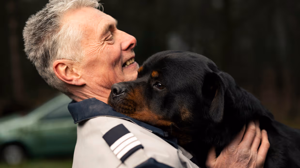 Links een man in uniform. Hij omarmt een grote zwart-bruine hond met opgetrokken oren. Achtergrond toont een bosrijke omgeving met een metalen hek. Rechts een glimlachende man in uniform staat naast een zwarte Rottweiler met open bek. Bosrijke omgeving op de achtergrond, met een ontspannen en vriendelijke sfeer. 