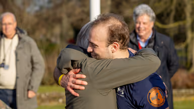 Links een groep mensen in militaire kleding loopt rennend over een keienpad, omringd door bomen en een gebouw. Aan de zijkant houden twee mannen een spandoek vast. Het is een zonnige dag. Rechts: twee mannen omarmen elkaar hartelijk buiten in de zon. De ene draagt een groene jas, de andere een donkerblauw sportshirt met een oranje logo. Op de achtergrond staan twee oudere mannen. 