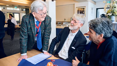 Een man in pak spreekt met twee heren die aan een tafel zitten in een zaal.