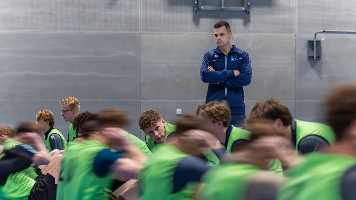 Jongeren in gele hesjes voeren diverse sportonderdelen uit in een sportzaal. 