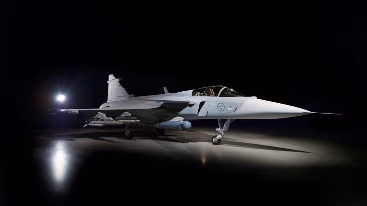 Een straaljager in een donkere hangar, verlicht door een felle lamp. Het vliegtuig staat stil op de grond, met een slanke, puntige neus en wapens onder de vleugels. 