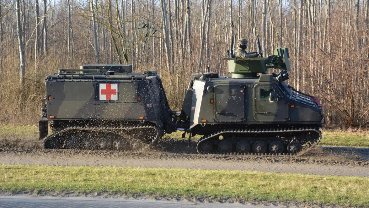 Een militair rupsvoertuig met een rood kruis op de zijkant, rijdend over onverhard terrein. Een soldaat zit bovenop, omgeven door bos.