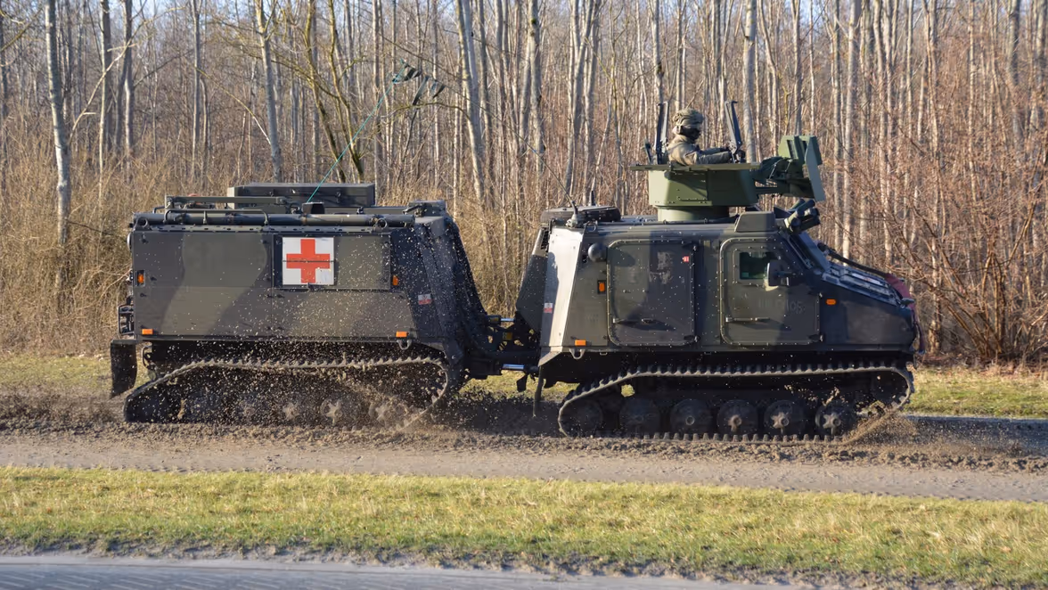 Een militair rupsvoertuig met een rood kruis op de zijkant, rijdend over onverhard terrein. Een soldaat zit bovenop, omgeven door bos.