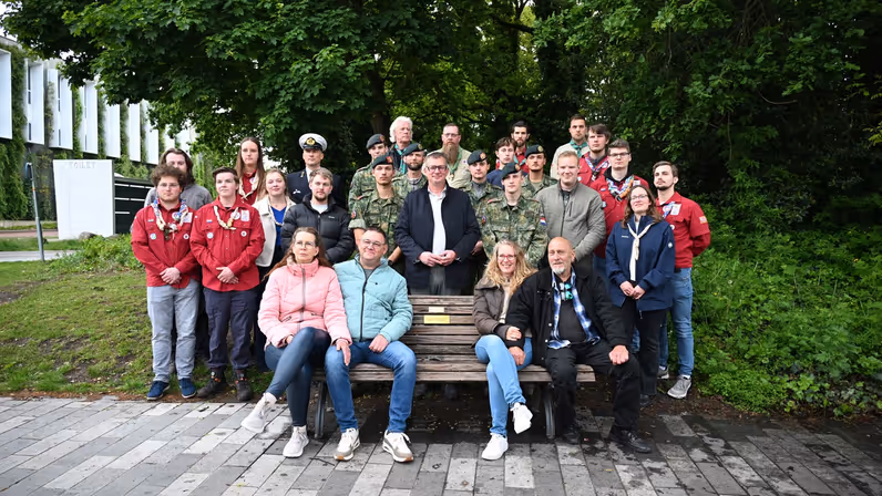 De (stief)ouders van Gijs op het bankje in Emmen met daarachter militairen, de wethouders en scouts. 