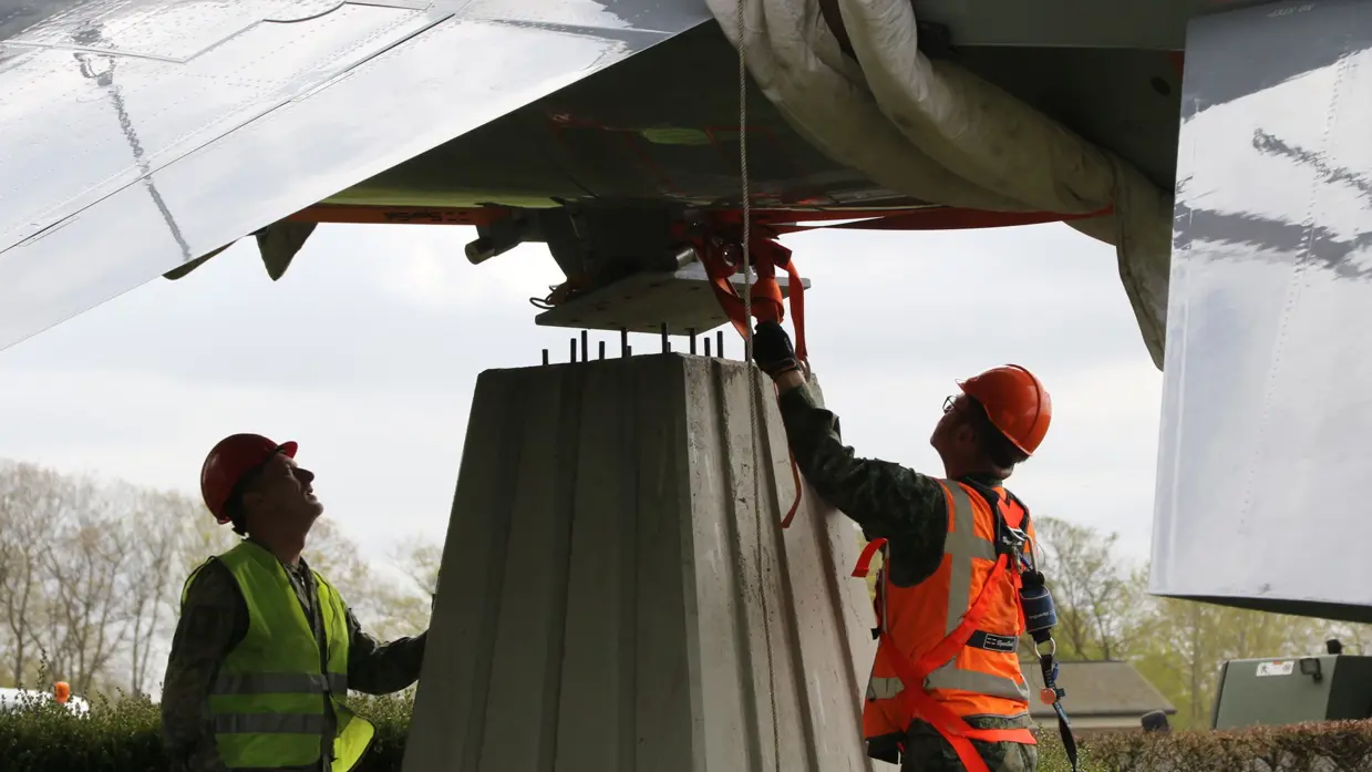 Twee arbeiders in veiligheidskleding bevestigen een groot metalen voorwerp onder de vleugel van een vliegtuig. 