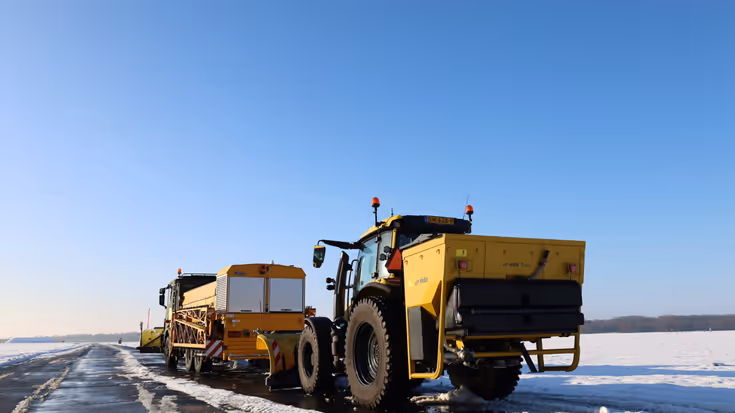 Gele sneeuwruimvoertuigen, waaronder een trekkers met sneeuwschuiver, werken op een besneeuwde startbaan onder een heldere, blauwe hemel.
