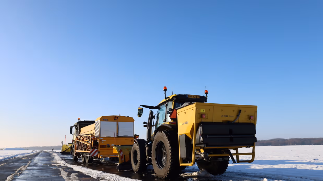 Gele sneeuwruimvoertuigen, waaronder een trekkers met sneeuwschuiver, werken op een besneeuwde startbaan onder een heldere, blauwe hemel.