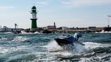 Een blauwe boot vaart met hoge snelheid over woest water, met een groen-witte vuurtoren en stad op de achtergrond.