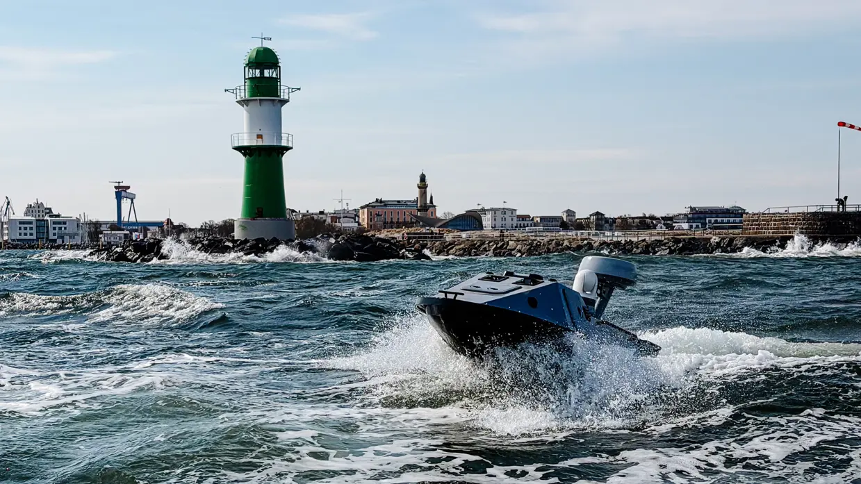 Een blauwe boot vaart met hoge snelheid over woest water, met een groen-witte vuurtoren en stad op de achtergrond.