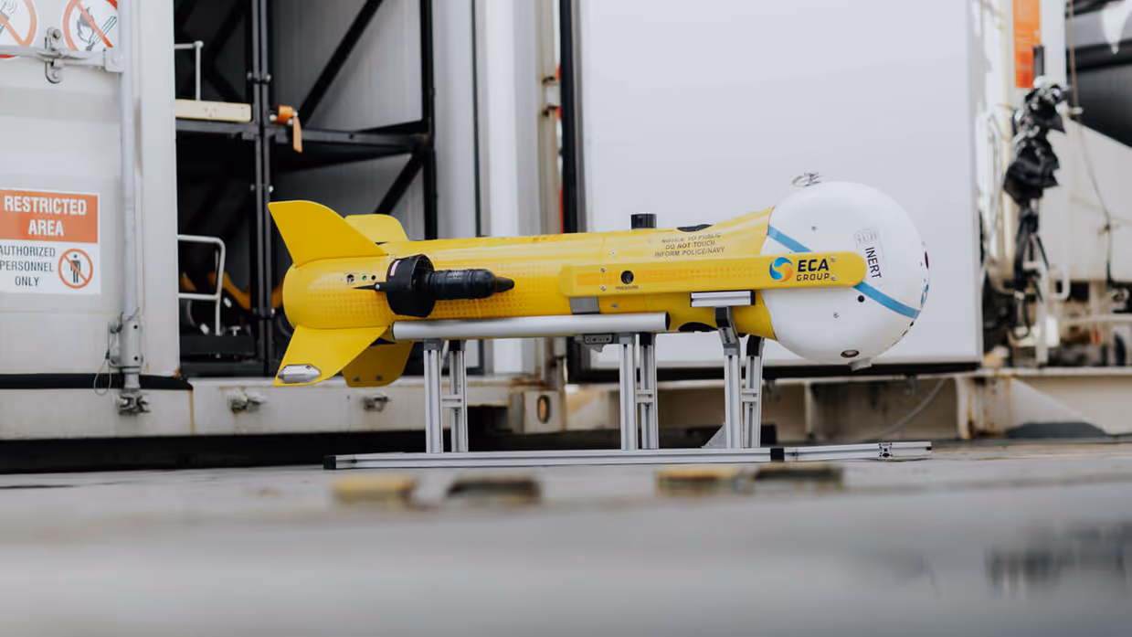 Links een grijs bootje met achterop verschillende drones. Dit vaart langs een groot vrachtschip. Daarnaast een gele, raketvormige drone met voorop een witte bol. Deze is ook te zien achterop het varende bootje.