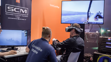 Een persoon in een t-shirt van marine praat in een gameroom tegen iemand in burgerkleding met VR-bril op.