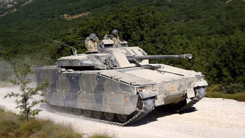  Een CV90-infanterievoertuig rijdt over de weg, een stofwolk achterlatend. Op de achtergrond zijn groene heuvels te zien.