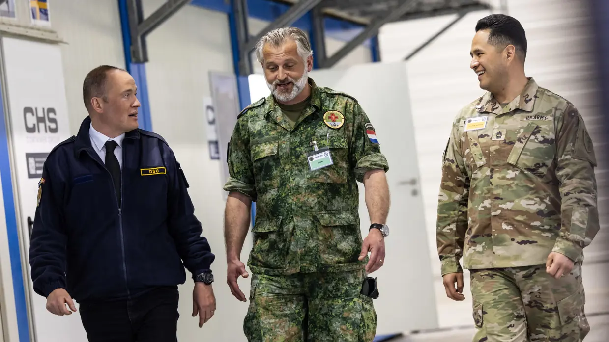 Links: Een man in pak met bril en identiteitskaart werkt op een laptop. Een vrouw in militair uniform met een medisch embleem staat naast hem. Rechts: drie mannen lopen glimlachend door een gang: een in donkerblauw uniform, een in Nederlands militair tenue met medisch embleem, en een in Amerikaans legeruniform. 