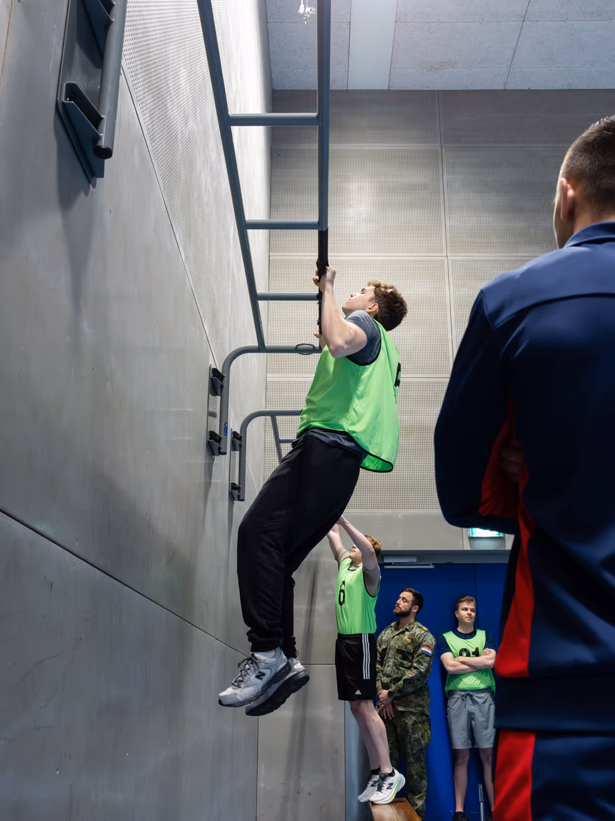 Jongeren in gele hesjes voeren diverse sportonderdelen uit in een sportzaal. 