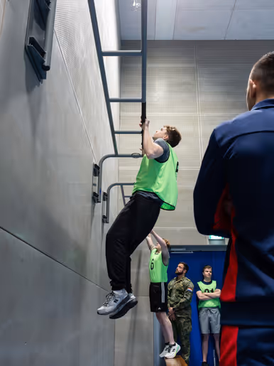 Jongeren in gele hesjes voeren diverse sportonderdelen uit in een sportzaal. 