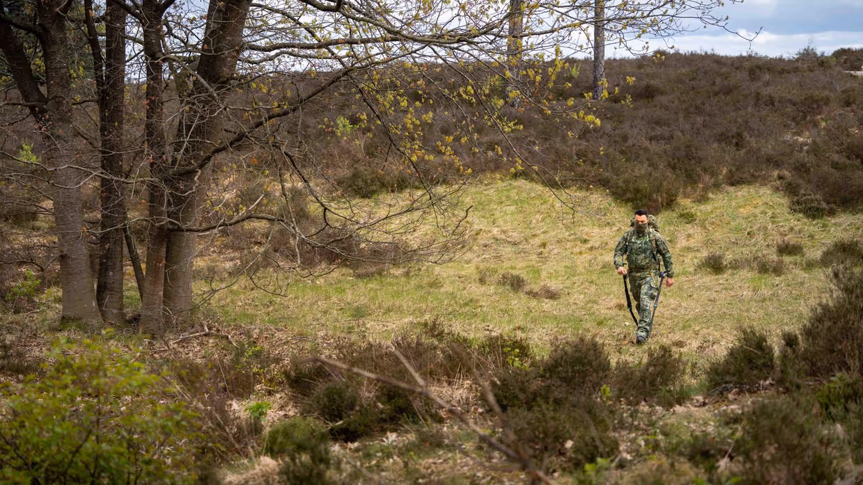 Een persoon in camouflage loopt met stokken door een bos met heide, bomen en groen gras onder een bewolkte lucht. 