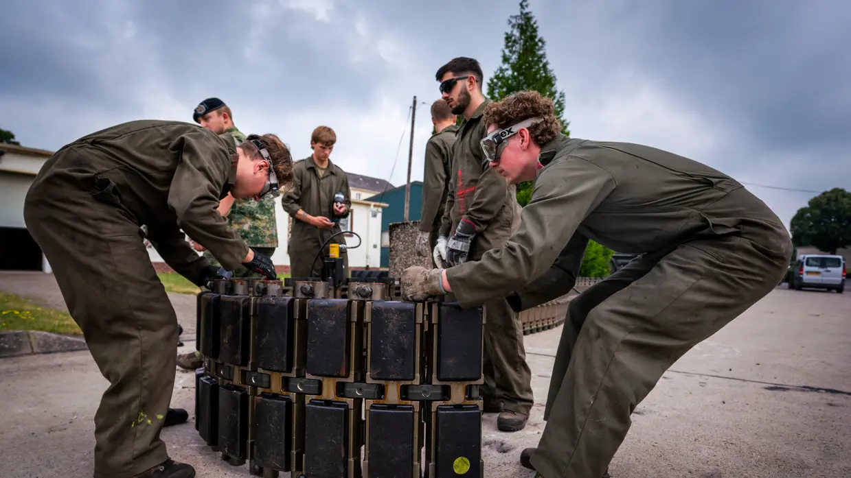 Handen in grijze werkhandschoenen draaien aan een metalen bout op een roestig, zwaar mechanisch onderdeel.