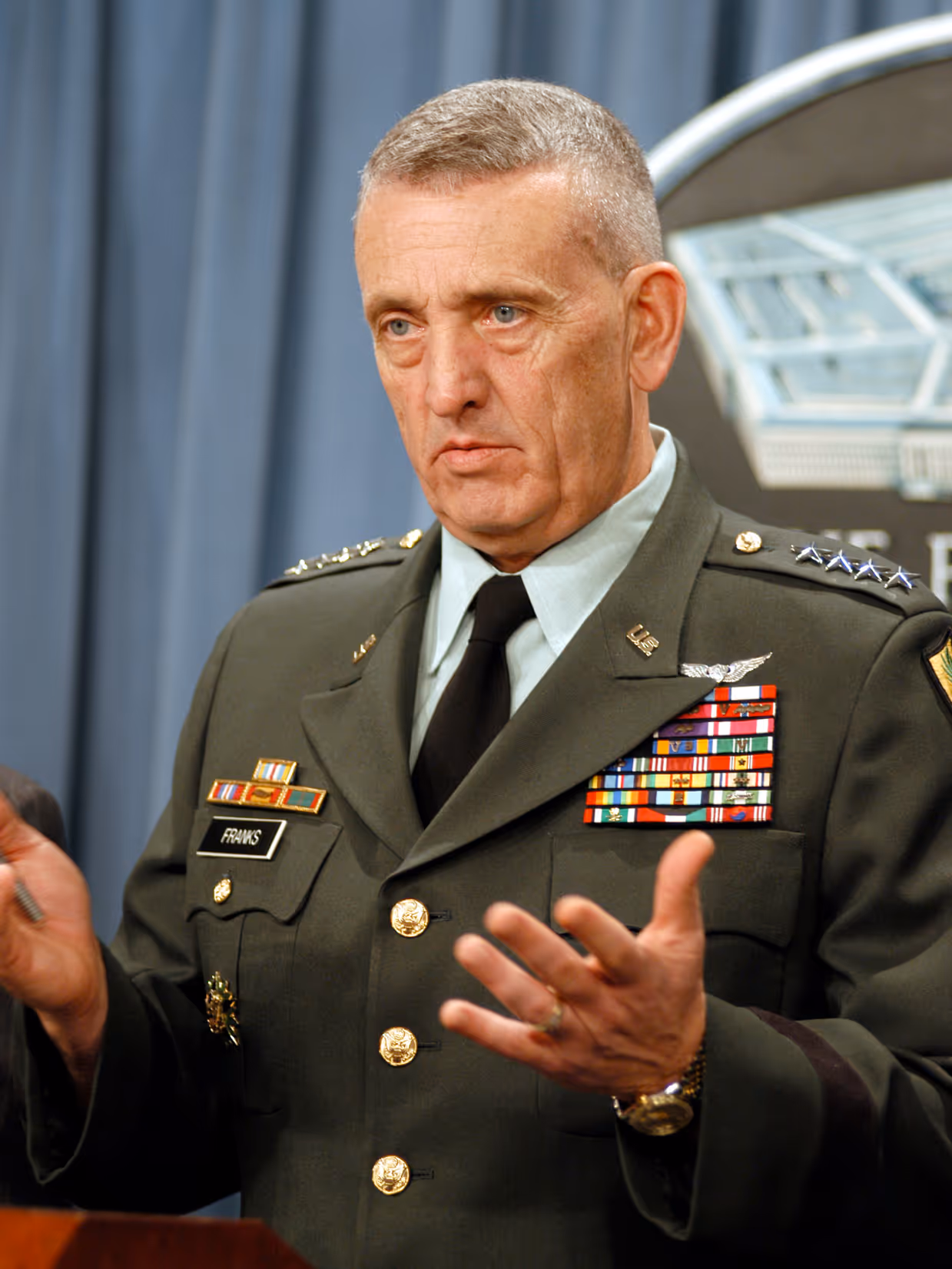 Een man in een groen militair uniform met veel medailles en sterren op zijn borst staat te praten met een ernstige blik. Hij heeft een naamplaatje met daarop ‘Franks’ op zijn uniform.