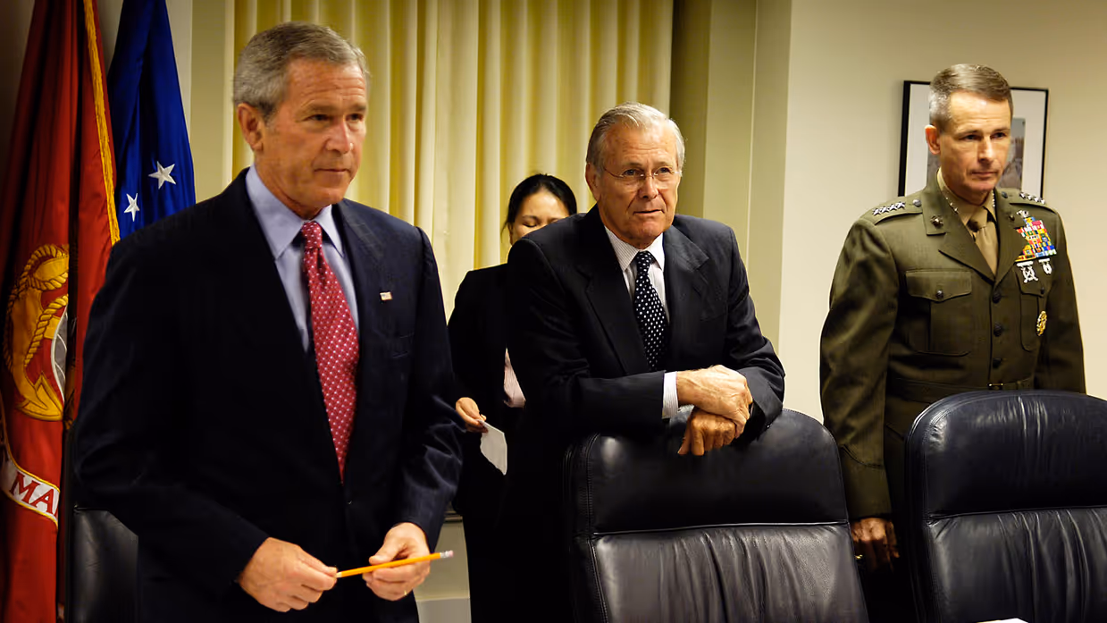 Drie mannen in pak, van wie een in uniform, staan achter stoelen aan een tafel met vlaggen op de achtergrond.