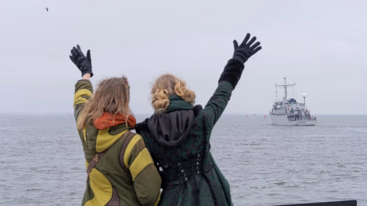 Twee mensen in warme kleding zwaaien enthousiast naar een grijs militair schip op kalme zee, terwijl een vogel boven hen vliegt.