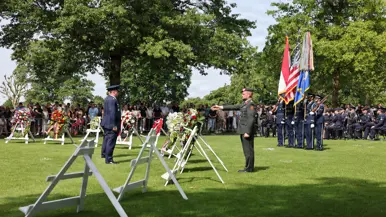 Een Amerikaanse militair staat in de houding tegenover een Nederlandse. In de achtergrond staan kransen en publiek. Rechts staan vier opgestelde militairen met vlaggen en zit het orkest van de luchtmacht.