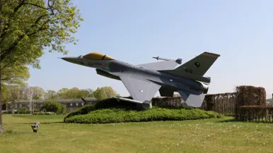 Een grijze straaljager staat schuin opgesteld in een groen park met bomen, struiken en een gebouw op de achtergrond. 