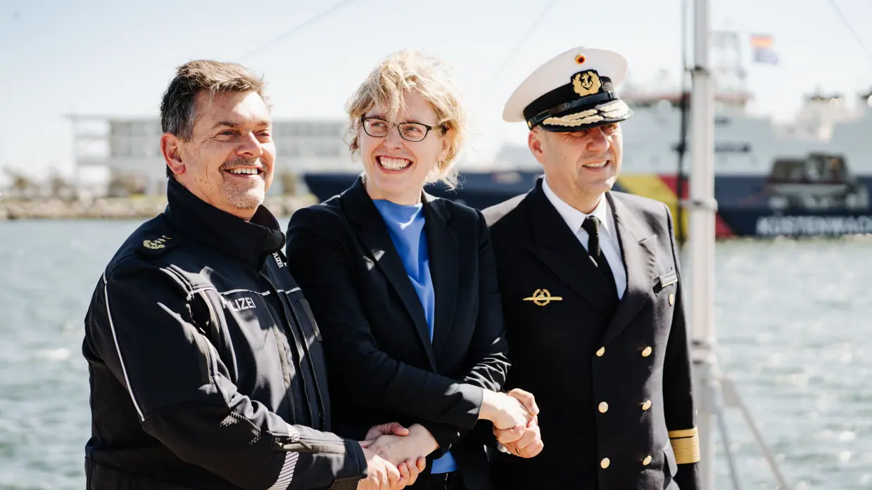 Drie mensen in uniformen schudden elkaar de hand op een schip, met water en een ander schip op de achtergrond.