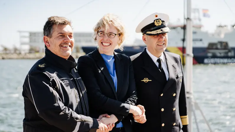 Drie mensen in uniformen schudden elkaar de hand op een schip, met water en een ander schip op de achtergrond.
