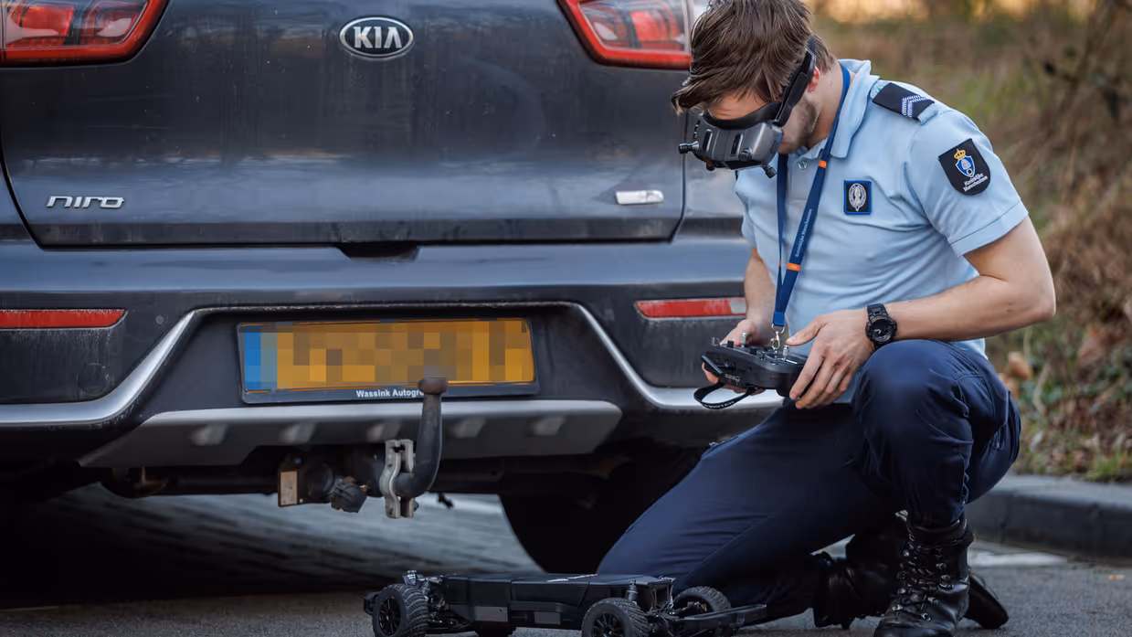 Een marechaussee bedient op een controller een robot die onder een auto verdwijnt. 