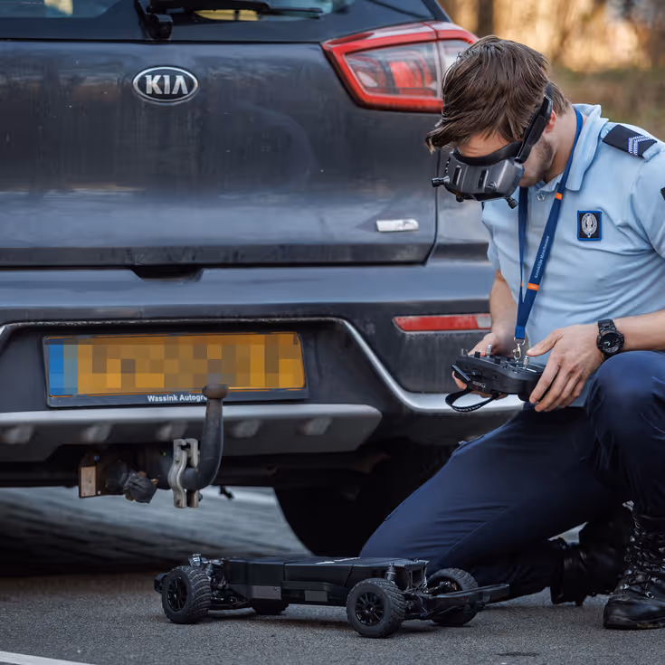 Een marechaussee bedient op een controller een robot die onder een auto verdwijnt. 