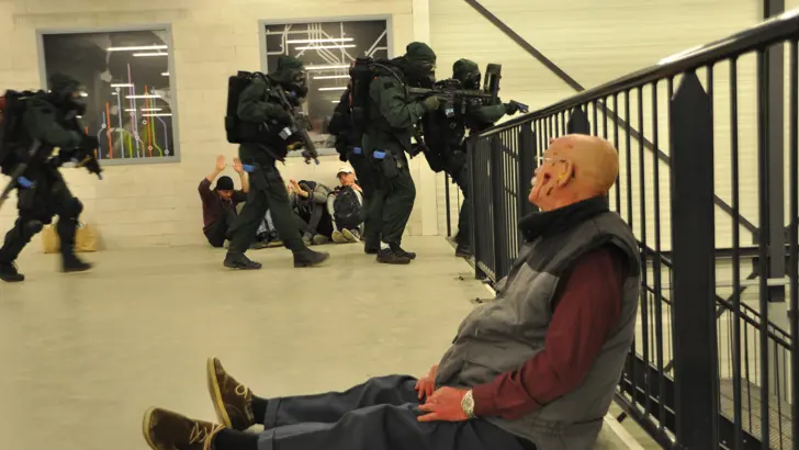 Gewonde man zit tegen een hek in een metrostation. Personen in beschermende pakken lopen een trap af.