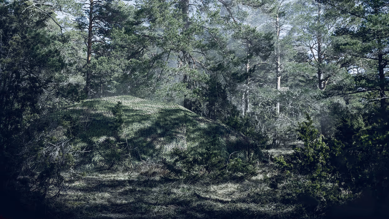 Een militair voertuig in het bos verstopt onder een groot camouflagenet.