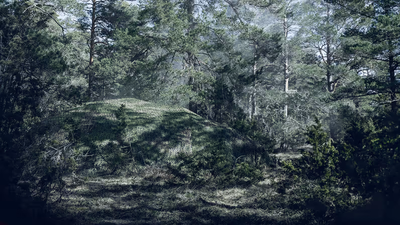 Een militair voertuig in het bos verstopt onder een groot camouflagenet.