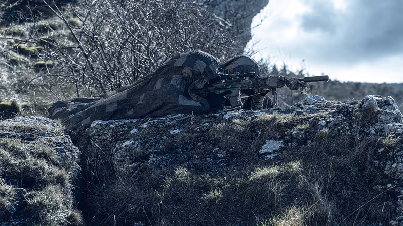 Een gewapende militair op een berg onder een camouflagedeken.