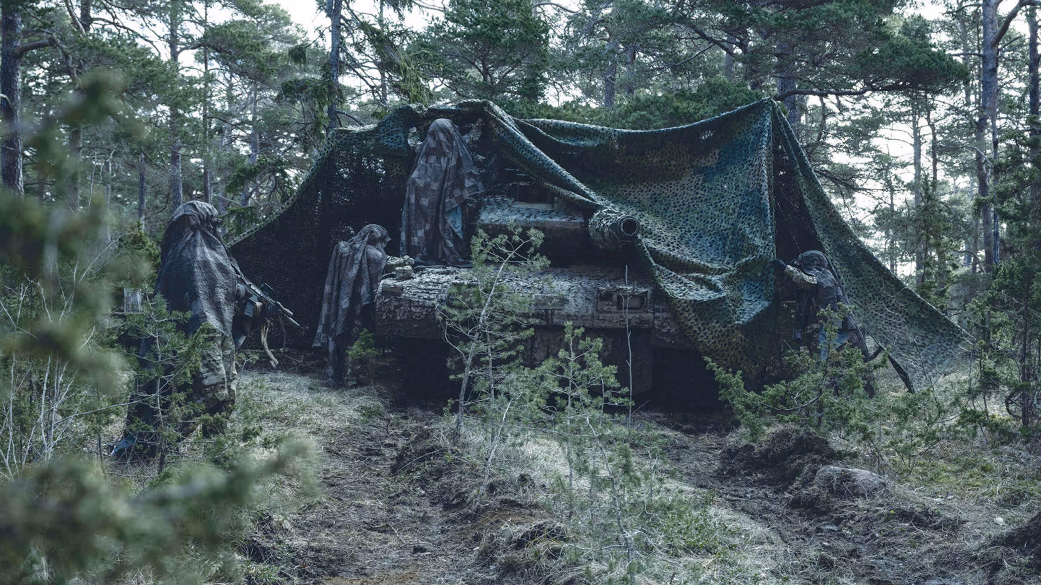 Militair voertuig in bos onder camouflagenet met daaromheen een aantal gecamoufleerde militairen.