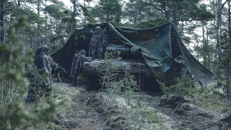 Militair voertuig in bos onder camouflagenet met daaromheen een aantal gecamoufleerde militairen.